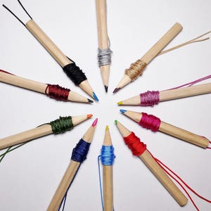 May include: A collection of wooden pencils with colorful string wrapped around the ends. The pencils are arranged in a starburst pattern on a white background.