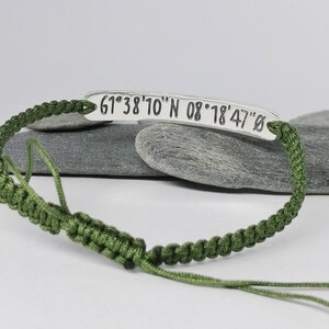 May include: A green cord bracelet with a silver tag that reads "61&deg;38'10" N 08&deg;18'47" O".