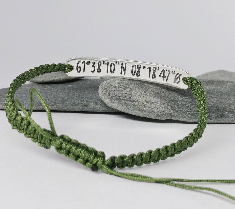 May include: A green cord bracelet with a silver tag that reads "61&deg;38'10" N 08&deg;18'47" O".