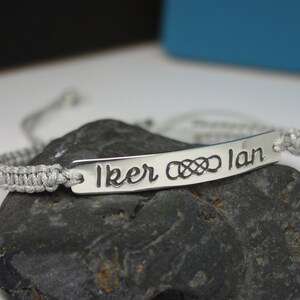 May include: A silver bracelet with a gray cord and a silver tag that says "Iker &infin; Ian".