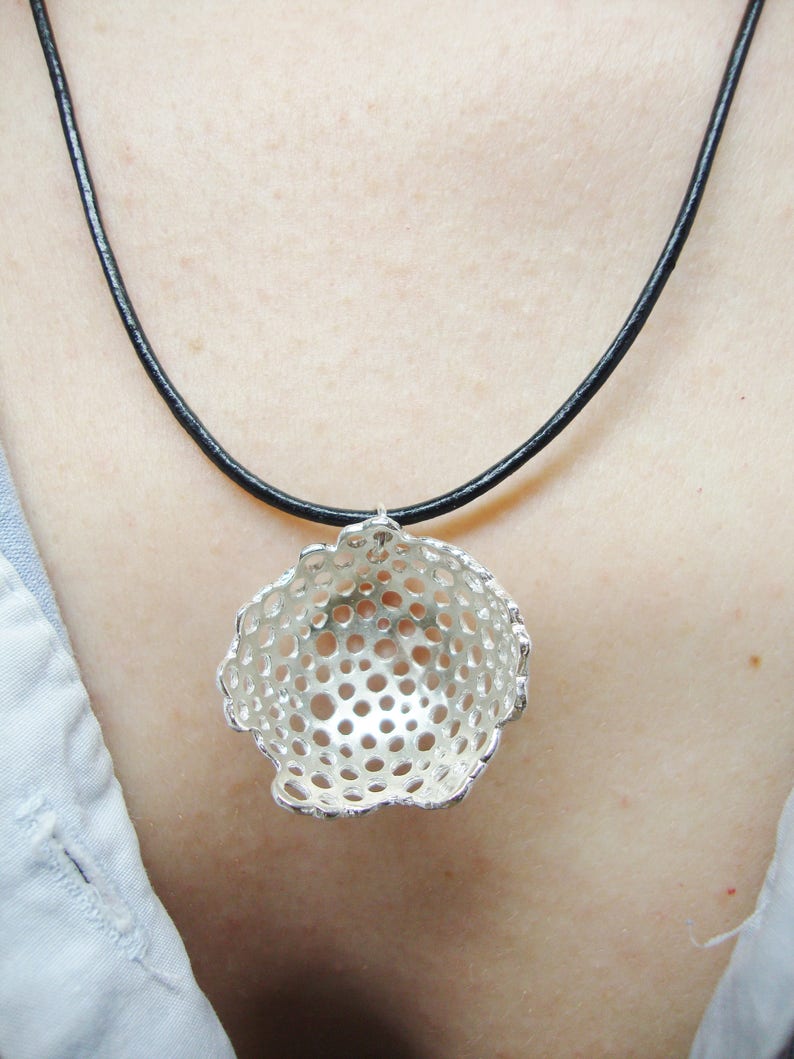 May include: A silver necklace with a delicate, openwork pendant shaped like a sphere. The pendant has a circular pattern of small holes throughout. The necklace is on a black cord.