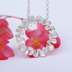 May include: A silver necklace with a circular pendant made of wavy, textured metal. The pendant is surrounded by pink flowers.
