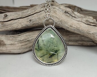 May include: A silver necklace featuring a large, pear-shaped pendant. The pendant has a light green, translucent stone with dark green inclusions, set in a beaded silver frame. The chain is a delicate link style.