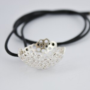 May include: A silver pendant necklace with a delicate, intricate design. The pendant is shaped like a small bowl with a honeycomb pattern. The necklace is on a black cord.