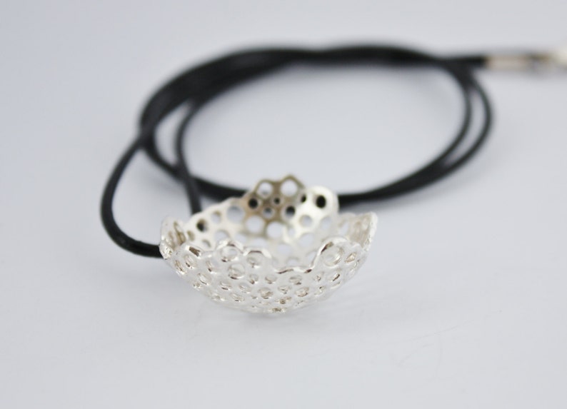 May include: A silver pendant necklace with a delicate, intricate design. The pendant is shaped like a small bowl with a honeycomb pattern. The necklace is on a black cord.