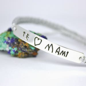May include: A silver bracelet with a gray braided cord and a silver charm with the words "Te amo Mami" engraved on it.