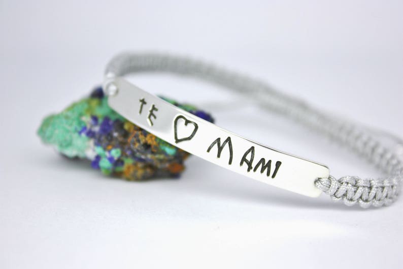 May include: A silver bracelet with a gray braided cord and a silver charm with the words "Te amo Mami" engraved on it.