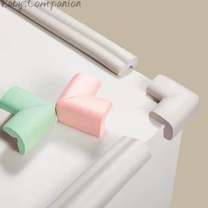 May include: A selection of colorful corner protectors in mint green, pink, and white. The image also shows white edge protectors. The text "BabysCompanion" is in the upper left corner. These are designed to protect children from sharp edges.