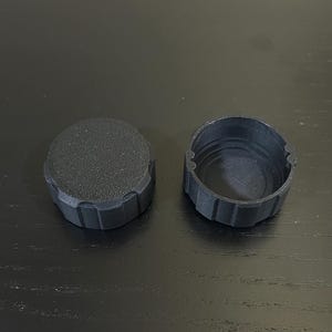 Könnte beinhalten: Zwei schwarze, 3D-gedruckte Kunststoffteile. Eines ist eine runde Kappe mit strukturierter Oberfläche und geriffelten Seiten. Das andere ist eine zylindrische Basis mit passendem geriffelten Design. Beide liegen auf einer dunklen Oberfläche.