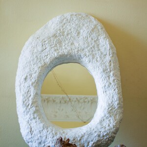 Puede incluir: Un espejo ovalado de color blanco con un marco grueso y texturizado que recuerda a la piedra. El espejo se apoya en la base sobre un trozo de madera flotante natural de color marrón. La superficie reflectante muestra el entorno.