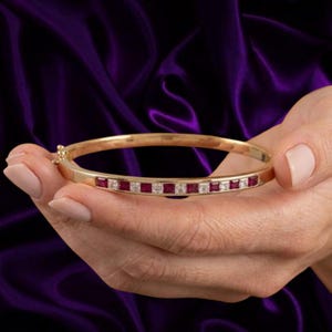 May include: A gold bangle bracelet featuring a row of alternating square-cut red rubies and clear white diamonds set into the metal band. The bracelet has a hinged clasp mechanism on the side and a polished finish throughout the gold surface.