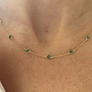Könnte beinhalten: Eine zarte Goldkette mit gleichmäßig verteilten, kleinen quadratischen grünen Edelsteinen in Goldfassungen. Die Kette liegt auf der Haut auf und präsentiert ein schlichtes Design mit einer dünnen Metallgliederkette und fünf sichtbaren Steinen.