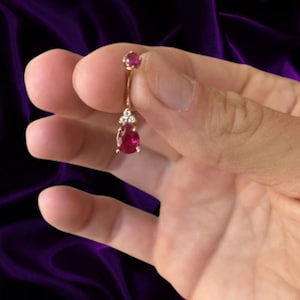 May include: A gold-toned navel piercing jewelry piece featuring a small round red gemstone at the top and a larger pear-shaped red gemstone at the bottom, accented by three tiny clear stones positioned between the two larger red gems.