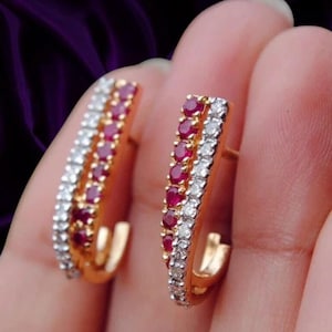 May include: A pair of gold hoop earrings featuring two parallel rows of gemstones. One row consists of small, round, deep red rubies, while the other row is set with small, round, clear white diamonds. The earrings have a curved, semi-circular shape.