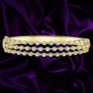 Może przedstawiać: Otwierana bransoletka typu bangle z żółtego złota z trzema równoległymi rzędami małych, okrągłych, przezroczystych kamieni ułożonych w ciągłej linii. Konstrukcja posiada polerowaną metalową ramę, która pewnie utrzymuje kamienie na całej długości.
