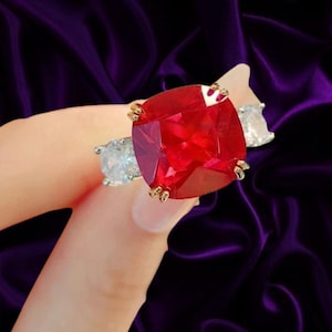 May include: A ring featuring a large, cushion-cut, vibrant red gemstone set in a gold-toned metal band. The central red stone is flanked on each side by a smaller, round, clear, faceted gemstone, creating a three-stone design with prong settings.