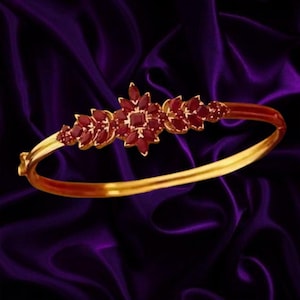 Puede incluir: Una pulsera rígida de oro con un grupo central de piedras preciosas de color rojo intenso, talladas en forma de marquesa y dispuestas en un patrón floral. La banda metálica es lisa y pulida, conectándose a la pieza central adornada.