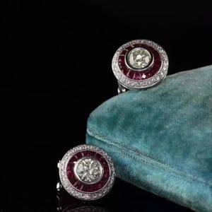 May include: A pair of circular earrings featuring a central round brilliant-cut diamond surrounded by a halo of deep red rubies, framed by an outer border of small pavé-set diamonds, all set in a polished white metal, resting on a teal velvet surface.