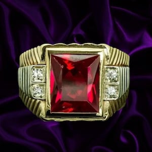 May include: A gold ring featuring a large, rectangular, deep red gemstone set in the centre. The band has a ribbed, textured design on both sides, with two small, round, clear, sparkling stones embedded vertically on each side of the centre gem.