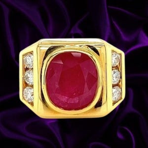 Puede incluir: Un anillo de oro con un gran rubí rojo intenso de corte ovalado en el centro. La banda está acentuada a ambos lados por una fila vertical de tres pequeños diamantes blancos redondos y claros, creando un diseño simétrico con acabado dorado.