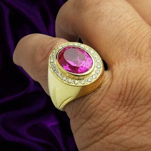 Puede incluir: Un anillo de oro con una gran piedra preciosa de color rosa vibrante y corte ovalado en el centro. La piedra central está rodeada por un halo de pequeñas piedras blancas de corte redondo engastadas en la banda de oro, que tiene un cuerpo ancho y pulido.