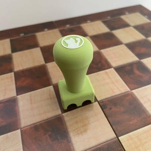 Puede incluir: Una pieza de ajedrez verde claro con un diseño de corona en la parte superior, colocada sobre un tablero de ajedrez de madera. La pieza tiene una parte superior redondeada y una base cuadrada con tres recortes arqueados. El tablero presenta cuadrados marrones claros y oscuros alternados.