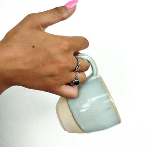 Puede incluir: Una taza de cerámica con una sección superior esmaltada en azul claro brillante y una base beige mate sin esmaltar. La taza tiene un asa curva y un cuerpo redondeado y liso, mostrando un acabado de dos tonos con variaciones sutiles.