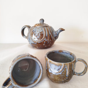 Può includere: Un set da tè in ceramica composto da una teiera e due tazze, caratterizzato da una smaltatura bicolore con base marrone lucido e un motivo a goccia azzurro. Gli oggetti presentano una superficie testurizzata con punti in rilievo.