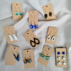 Puede incluir: Una colección de diez pares de pendientes de cerámica presentados en tarjetas de cartón marrón. Los pendientes tienen varias formas, incluyendo estrellas, gotas y figuras geométricas en colores como azul, verde, blanco, negro y moteados.