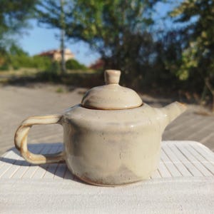 Puede incluir: Tetera de cerámica beige con tapa, boquilla y asa. El asa tiene un diseño retorcido. La tetera está sobre una superficie blanca, con un fondo borroso de árboles y cielo. La tetera mide aproximadamente 15 cm de largo.