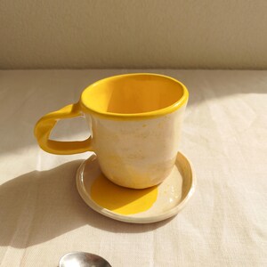Pode incluir: Um conjunto de chávena e pires em cerâmica artesanal com interior e rebordo amarelo brilhante e exterior creme com salpicos amarelos. A chávena tem uma pega grossa e curva, e uma pequena colher de metal prateado repousa ao lado.