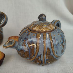 Può includere: Piccola teiera in ceramica con smalto blu e marrone. La teiera ha un corpo arrotondato, un coperchio con un piccolo pomello e un beccuccio curvo. Il design presenta strisce marroni verticali e piccoli punti. Un'aggiunta affascinante a qualsiasi set da tè.