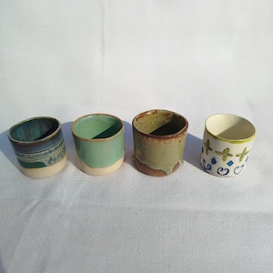 Puede incluir: Cuatro tazas pequeñas de cerámica dispuestas en fila. La primera tiene un esmalte azul y crema. La segunda es verde claro con base crema. La tercera es de color verde oliva y marrón moteado. La cuarta es blanca con un patrón floral azul y verde.