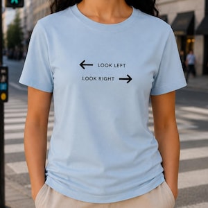 Könnte beinhalten: Ein hellblaues T-Shirt mit kurzen Ärmeln und schwarzem Text sowie Grafiken auf der Brust. Das Design zeigt einen nach links zeigenden Pfeil mit dem Text LOOK LEFT über einem nach rechts zeigenden Pfeil mit dem Text LOOK RIGHT.