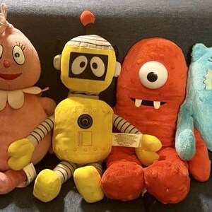 May include: A set of four plush toys including a pink round character with a white collar, a yellow robot with striped arms and an antenna, a textured orange one-eyed monster with two white teeth, and a blue furry creature with two teeth.