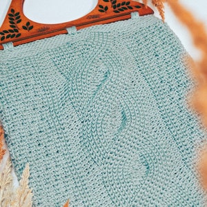 Puede incluir: Una bolsa de mano de crochet azul claro con un asa de madera con un diseño de hojas talladas.