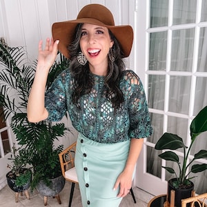 May include: A woman wearing a green and blue crochet sweater with a light blue button-down skirt and a brown wide-brimmed hat.
