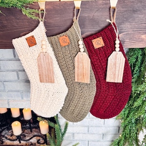 Könnte beinhalten: Drei gehäkelte Weihnachtsstrümpfe hängen an einem Holzmantel. Die Strümpfe sind weiß, grün und rot. Jeder Strumpf hat ein Holzschild mit einem Ledermonogramm.