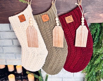 May include: Three crocheted Christmas stockings in cream, olive green, and deep red. Each stocking features a brown leather patch with a geometric logo, a jute hanging loop, and a wooden gift tag suspended by a string of three small wooden beads.