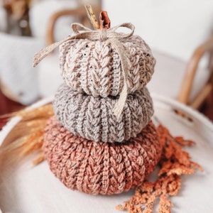 A stack of three knitted pumpkins in shades of beige, gray, and rust is displayed on a wooden tray, adorned with a ribbon and surrounded by dried orange foliage, making it the perfect handmade gift for autumn lovers.