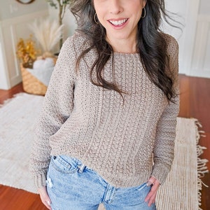 May include: A woman wearing a light brown cable knit sweater with long sleeves and blue jeans.