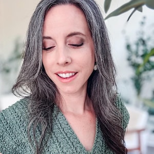 May include: A sage green short-sleeved top featuring a textured, vertical knit pattern and a V-neckline. The garment is constructed with a thick, yarn-based material that creates a distinct ribbed appearance across the front and sleeves.