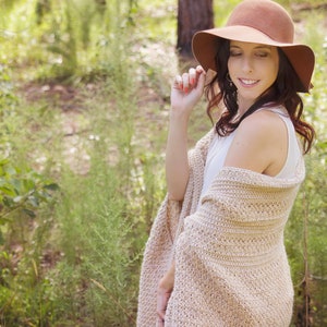 May include: A woman wearing a brown wide-brimmed hat and a beige crochet shawl, standing in a green grassy area.