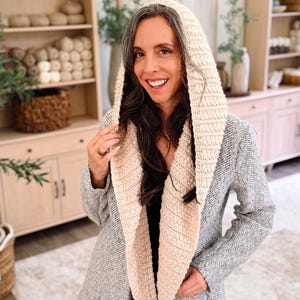 May include: A cream-colored crochet hood with a textured, ribbed stitch pattern. The hood is designed to be worn over the head and drapes down the front of the chest, featuring a wide, thick border that frames the face with a consistent knit texture.