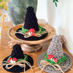 May include: Three crocheted hats in black and gray, each featuring a pointed crown and a wide brim. The hats are decorated with green moss, small red mushrooms with white spots, and a tied piece of natural tan twine at the base of the crown.
