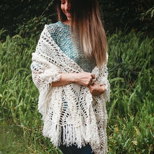 A woman wearing a white shawl decorated with the intricate crochet pineapple stitch, with fringed edges. She stands in an outdoor green space, surrounded by greenery and sunshine.