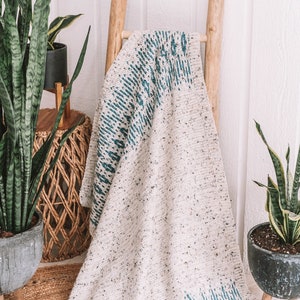 Puede incluir: Una manta de crochet blanca y azul con un patrón geométrico drapeada sobre una escalera de madera. La manta está sentada sobre una alfombra tejida frente a una pared blanca.