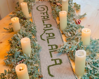 May include: A crocheted table runner with the word "Christmas" in green yarn on a beige background. The runner is decorated with a garland of green leaves and white candles.