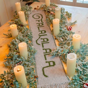 May include: A crocheted table runner with the word "Christmas" in green yarn on a beige background. The runner is decorated with a garland of green leaves and white candles.