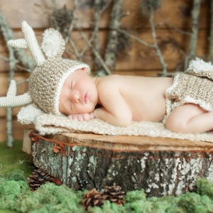 May include: A newborn baby wearing a crocheted deer hat and matching diaper cover sleeps on a tree stump covered in moss.